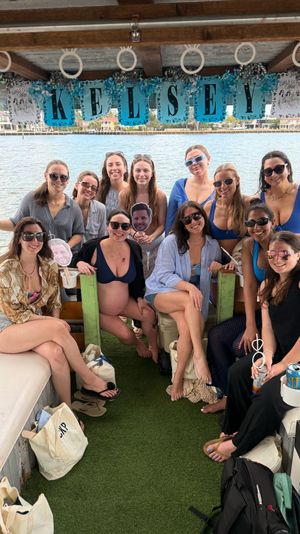 Group of smiling women on a decorated daytime party boat with waterfront homes in the background, personalized banner overhead, tote bags and party props, one visibly pregnant guest.