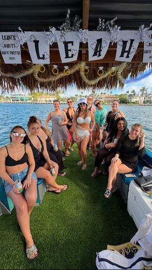 Group of women in swimsuits on a thatched-roof party boat with artificial turf, holding canned drinks under a decorated bachelorette banner, cruising past waterfront homes and palm trees on a sunny day