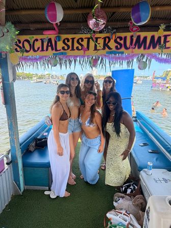 Six friends in swimsuits smiling and posing under a colorful "Social Sister Summer" tiki canopy on a floating boat bar, with other boats and a sunny coastal bay in the background.