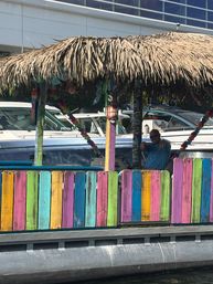 Vibrant tiki hut with a thatched roof and rainbow-painted fence on a marina dock, boats behind and a person waving inside.
