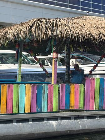 Vibrant tiki hut with a thatched roof and rainbow-painted fence on a marina dock, boats behind and a person waving inside.