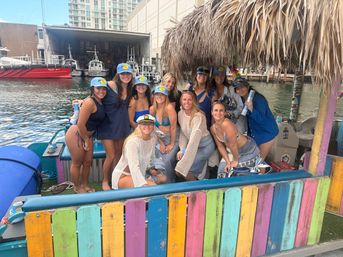Group of smiling women in swimsuits and matching caps posing on a colorful tiki-roofed boat at a city marina, with pilot boats and waterfront buildings in the background.