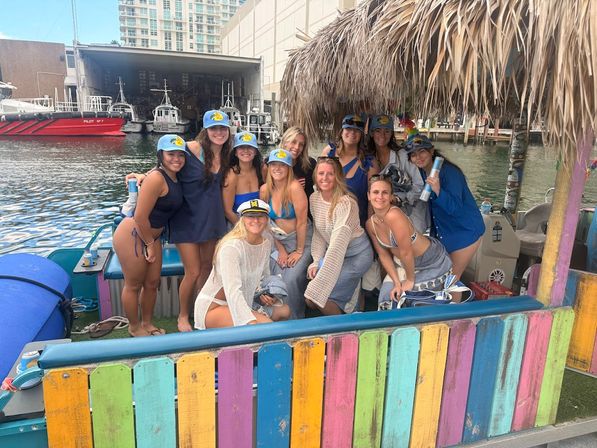 Group of smiling women in swimsuits and matching caps posing on a colorful tiki-roofed boat at a city marina, with pilot boats and waterfront buildings in the background.