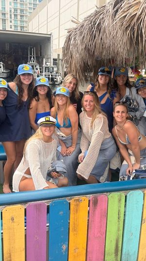 Group of smiling women in swimsuits and matching caps posing on a colorful tiki-roofed boat at a city marina, with pilot boats and waterfront buildings in the background.