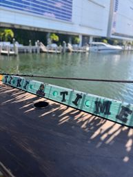 Turquoise wooden 'Tiki Time' sign on a boat railing overlooking a sunny waterfront marina with yachts, docks, and a modern waterside building