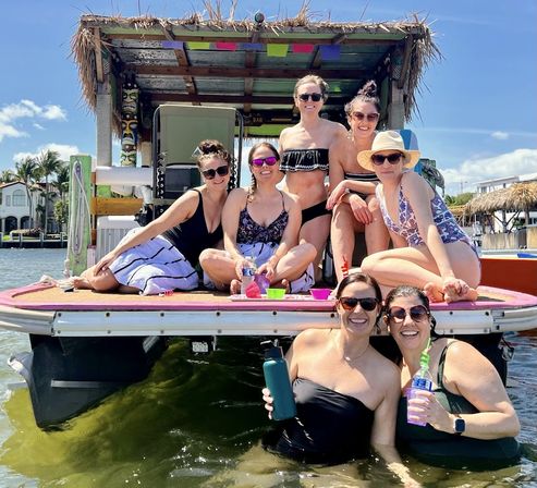 Seven women in swimwear smiling and holding drinks on a tikiâroof pontoon boat at a sunny waterfront bay, some wading in the water.