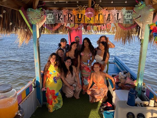 Group of friends posing on a colorful tiki-style boat over the water, tropical decorations and a happy birthday banner, women in swimsuits and summer dresses holding drinks at sunset
