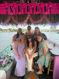 Six friends posing on a decorated waterfront tiki boat for a bachelorette — hot pink 'SARAH' banner overhead, bride in white holding a 'BRIDE' clutch, tropical cushions and a city skyline across the water.