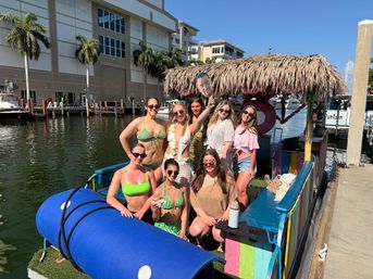 Eight women in swimsuits posing on a colorful tiki-style pontoon boat at a sunny waterfront marina with palm trees and docked boats, one holding a cardboard face cutout.