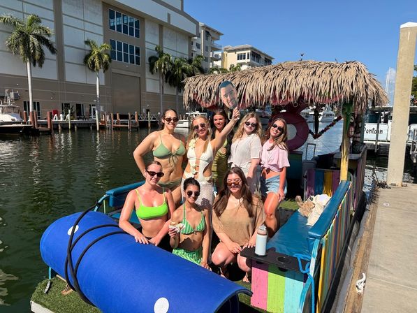Eight women in swimsuits posing on a colorful tiki-style pontoon boat at a sunny waterfront marina with palm trees and docked boats, one holding a cardboard face cutout.