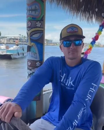 Man in blue fishing shirt and cap steering a tiki-decorated boat with mirrored sunglasses, colorful lei, and a coastal marina with boats in the background