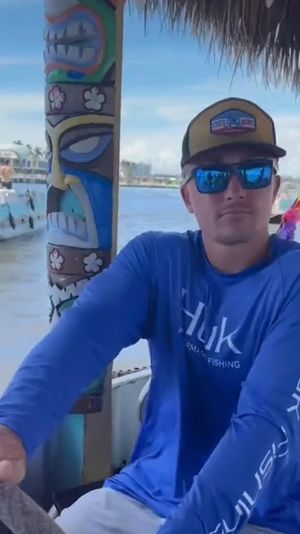 Man in blue fishing shirt and cap steering a tiki-decorated boat with mirrored sunglasses, colorful lei, and a coastal marina with boats in the background