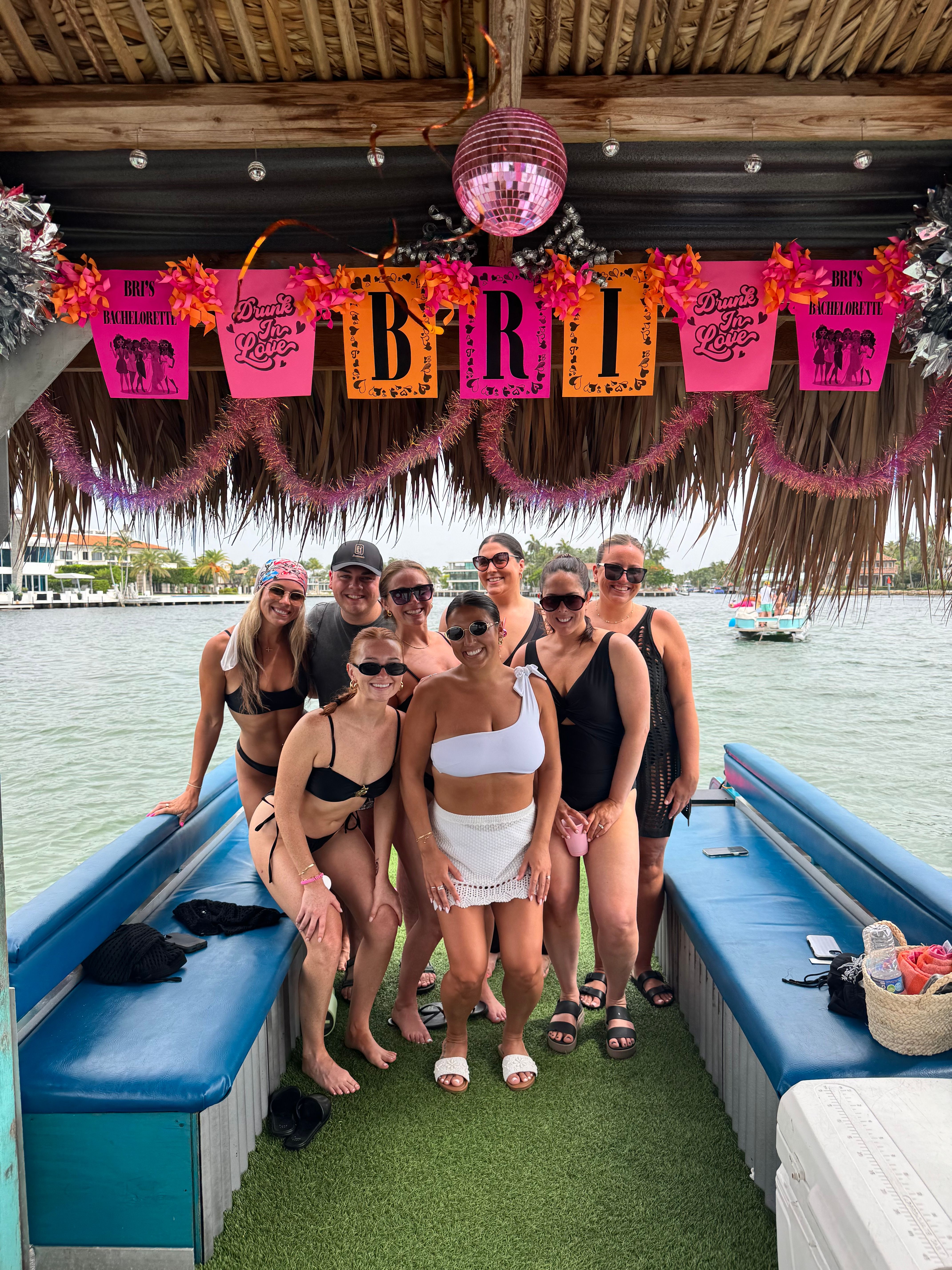 Eight friends in swimsuits posing on a tiki-boat deck by the water under pink bachelorette banners, tinsel and a disco ball with a palm-lined shoreline in the background.