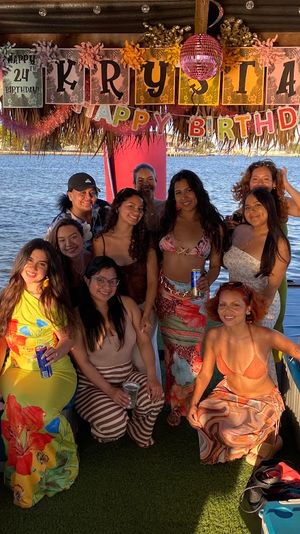 Group of friends in summer outfits posing on a colorful thatched-roof party boat at sunset on a lake, with a birthday banner, disco ball and drinks visible.
