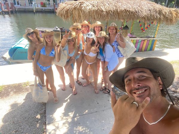Group of friends in bikinis and straw hats posing for a selfie by a tropical waterfront tiki hut and dock with a pontoon boat in the background