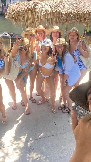 Group of friends in bikinis and straw hats posing for a selfie by a tropical waterfront tiki hut and dock with a pontoon boat in the background