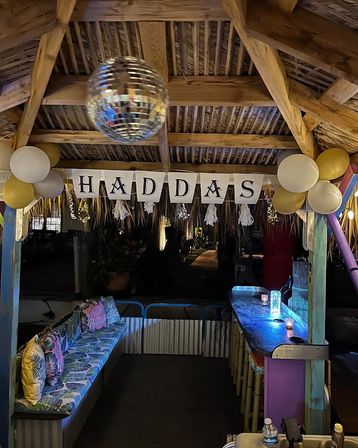 Backyard tiki bar at night decorated for a celebration with wooden rafters, hanging disco ball, gold-and-white balloons, âHADDASâ banner, tropical cushions and a glowing bar.