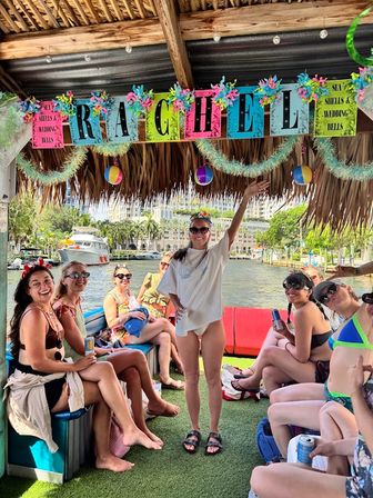 Group of friends on a decorated tiki-style party boat at a sunny waterfront marina — women in swimsuits holding drinks under colorful beach-themed banners, garlands and hanging beach balls.