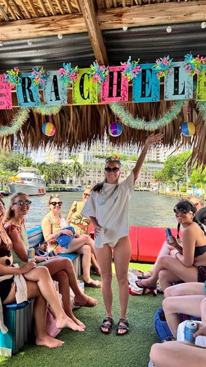 Group of friends on a decorated tiki-style party boat at a sunny waterfront marina — women in swimsuits holding drinks under colorful beach-themed banners, garlands and hanging beach balls.
