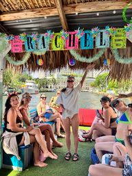 Group of women in swimsuits smiling on a decorated tiki-style boat under a colorful “RACHEL” banner, cruising a palm-lined waterfront canal