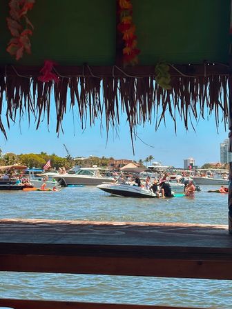 Tiki-thatched roof framing a sunny waterfront scene with anchored boats, people swimming and paddleboarding in a busy coastal bay with palm trees and buildings in the background.