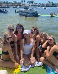 Seven women in swimsuits smiling on a green floating mat at a sunny waterfront boat party, holding canned drinks and wearing sunglasses and a white cap, matching circular arm tattoos visible, pontoon boats and palm-lined shoreline in the background.