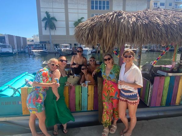 Group of seven women in colorful tropical outfits smiling with drinks at a thatched-roof tiki bar on a sunny marina dock, colorful painted wood panels and boats in the background.