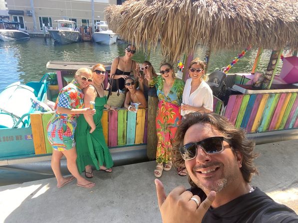 Smiling group of friends on a colorful tiki-style boat at a sunny marina dock, holding drinks while a man in the foreground takes a shaka selfie.