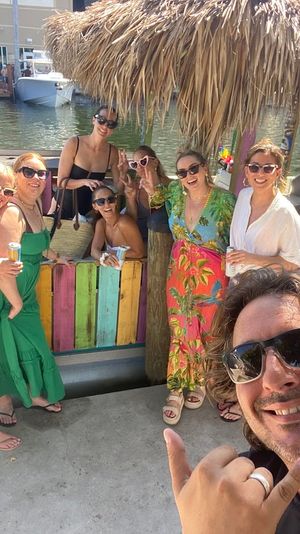 Smiling group of friends on a colorful tiki-style boat at a sunny marina dock, holding drinks while a man in the foreground takes a shaka selfie.