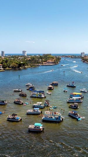 Aerial view of a sunny coastal inlet with dozens of party boats and people on inflatables clustered in the water, waterfront homes and high-rise buildings along the shore and the blue ocean on the horizon.