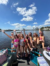 Group of women in colorful bikinis and heart-shaped sunglasses cheering on a pontoon boat under a bright blue sky over a palm-lined waterfront.