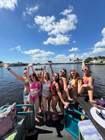 Group of women in colorful bikinis and heart-shaped sunglasses cheering on a pontoon boat under a bright blue sky over a palm-lined waterfront.