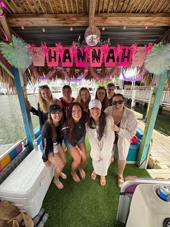 Group of smiling friends on a tiki-style party boat at a marina dock, decorated with pink party banner, disco ball, tinsel and coolers on an artificial grass waterfront deck.