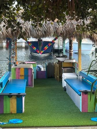 Colorful tiki-style dock at a tropical marina with a thatched roof, bright painted benches on artificial turf and a lei-decorated boat moored nearby