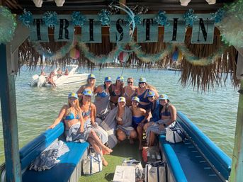 Group of friends in bikinis wearing matching caps on a decorated tiki-style party boat in shallow green bay with beach balls and garlands overhead