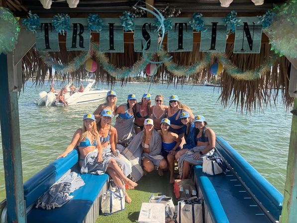 Group of friends in bikinis wearing matching caps on a decorated tiki-style party boat in shallow green bay with beach balls and garlands overhead