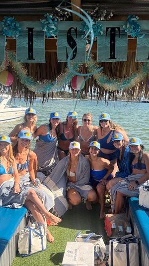 Group of friends in bikinis wearing matching caps on a decorated tiki-style party boat in shallow green bay with beach balls and garlands overhead