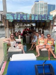 Smiling group of friends relaxing on a colorful tiki-themed party boat docked at a waterfront marina with high-rise condos and a waterway in the background.