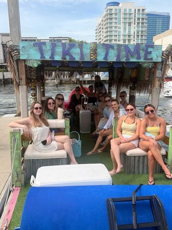 Smiling group of friends relaxing on a colorful tiki-themed party boat docked at a waterfront marina with high-rise condos and a waterway in the background.