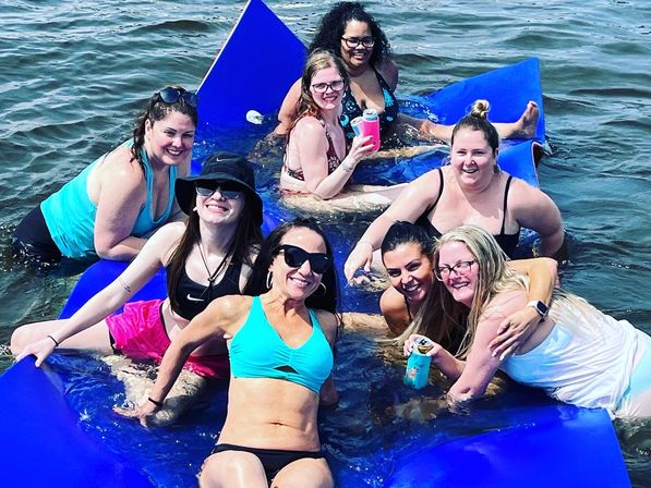 Group of eight women in swimsuits lounging on large blue floating mats in a lake, smiling and holding canned drinks during a sunny summer lake outing