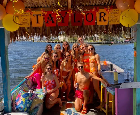 Group of friends in colorful swimsuits posing under a thatched tiki-roof on a sunny waterfront boat party with balloons, tropical cushions and a marina in the background.