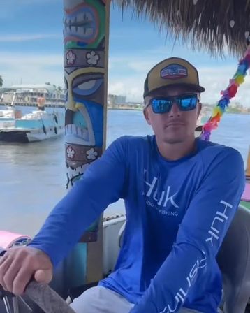 Person steering a colorful tiki-style boat in a sunny coastal marina, wearing a blue fishing shirt, mirrored sunglasses and cap with boats and waterfront buildings in the background.