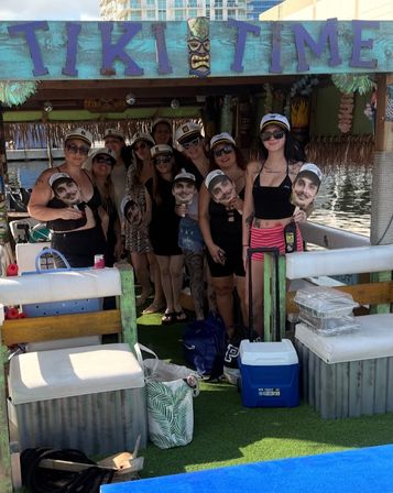 Group of friends posing on a tiki-style party boat at the dock, wearing captain hats and sunglasses and holding oversized face cutouts amid coolers, bags and tropical decor.