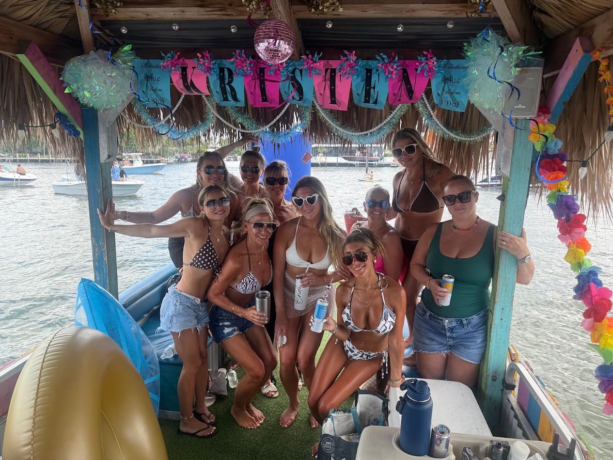 Group of women in swimsuits posing and smiling under a colorful thatched tiki hut on a party boat in a sunny marina, holding drinks with inflatables and other boats visible on the water — festive tropical boat celebration.