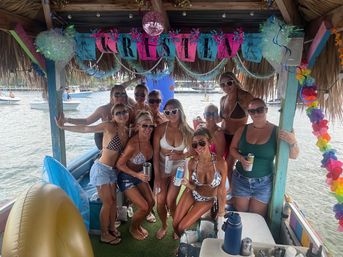 Group of women in swimsuits posing and smiling under a colorful thatched tiki hut on a party boat in a sunny marina, holding drinks with inflatables and other boats visible on the water — festive tropical boat celebration.