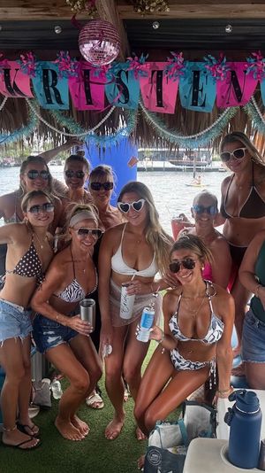 Group of women in swimsuits posing and smiling under a colorful thatched tiki hut on a party boat in a sunny marina, holding drinks with inflatables and other boats visible on the water — festive tropical boat celebration.