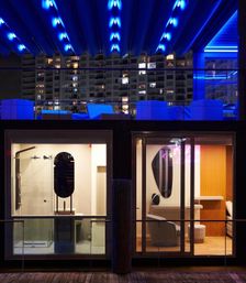 Nighttime urban waterfront scene: two glass-front modern rooms above a wooden dock with a rooftop lounge bathed in electric blue LED lights and high-rise apartments in the background.