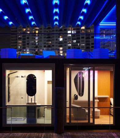 Nighttime urban waterfront scene: two glass-front modern rooms above a wooden dock with a rooftop lounge bathed in electric blue LED lights and high-rise apartments in the background.