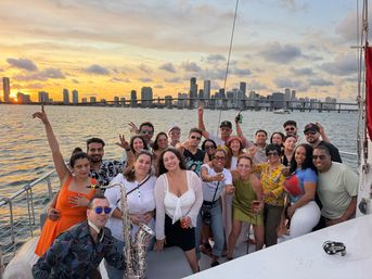 Smiling group of friends partying on a sailboat at sunset, drinks and a saxophone visible, with the Miami skyline and bridge over Biscayne Bay in the background