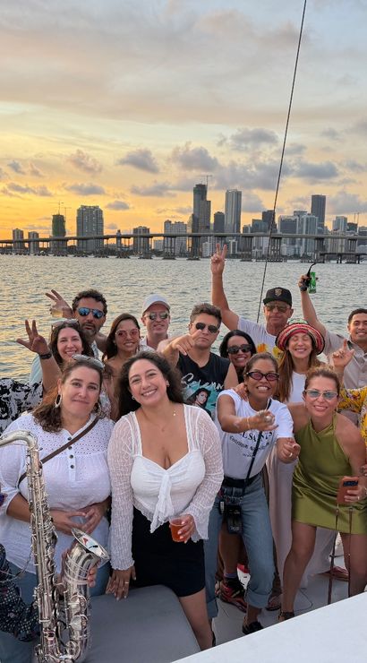 Smiling group of friends partying on a sailboat at sunset, drinks and a saxophone visible, with the Miami skyline and bridge over Biscayne Bay in the background